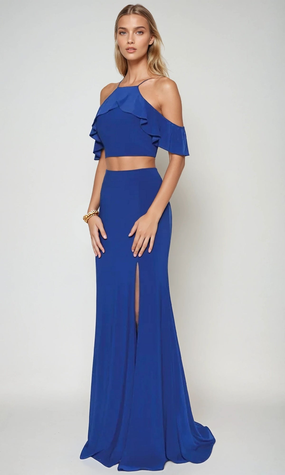 A woman wearing the Juliet 662 long prom dress—a royal blue two-piece with a cold-shoulder ruffled crop top and a high-waisted, floor-length skirt featuring a bold leg slit.